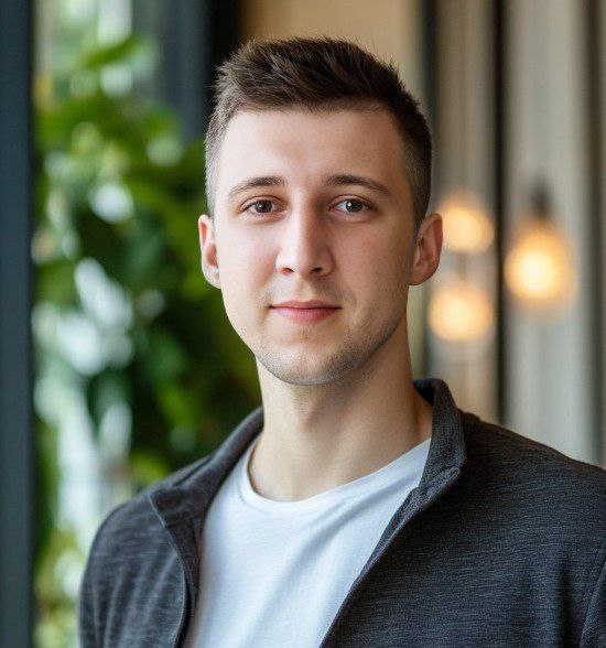 Young man with short brown hair wearing a white shirt and dark jacket standing indoors with blurred green plants and lights in the background.