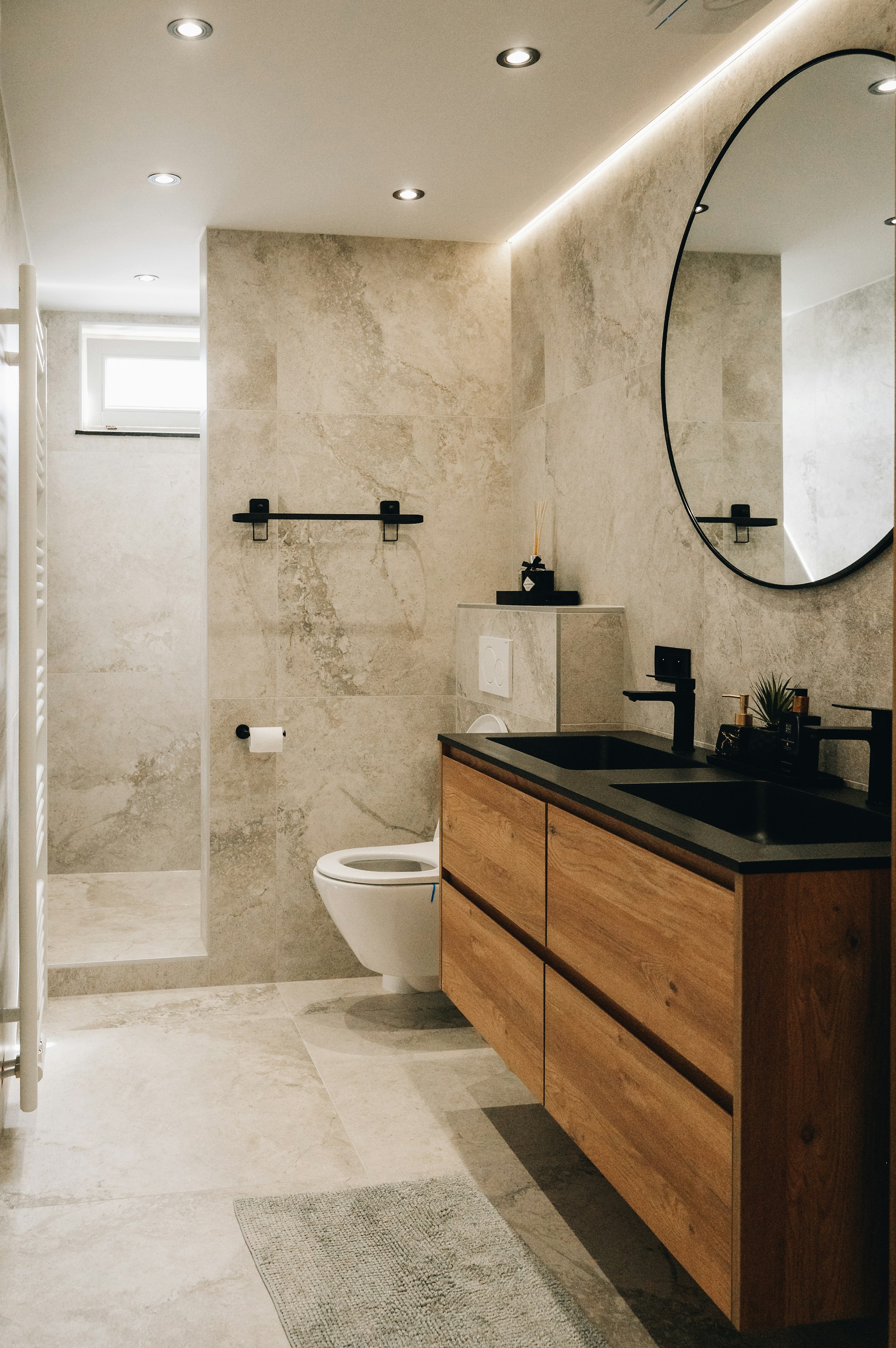 Modern bathroom with beige stone walls and floor, floating wooden vanity with black countertop and double sinks, round mirror, wall-mounted toilet, and walk-in shower.