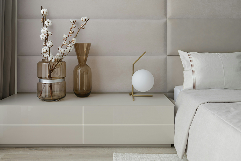 Modern beige bedside table with two brown translucent vases holding cotton stems and a gold table lamp with a white spherical shade, next to a bed with white and beige bedding.